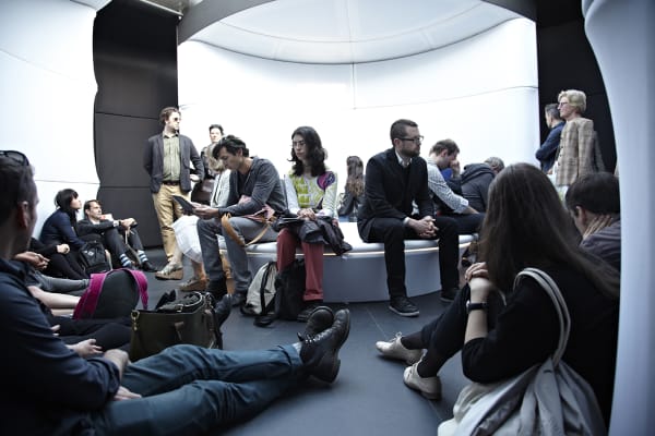 A group of people sat in a circular room with white walls.  Some are sat on a circular seat in the centre and others are sat on the floor.