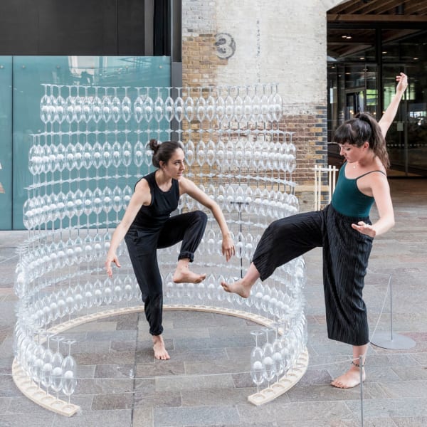 Two people mimicking each other's balance surrounded by a semi circular tower of wine glasses