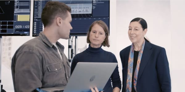 A man wearing a grey corduroy shirt, a woman wearing a black turtleneck and a woman wearing a dark navy blazer with a colourful silk scarf, standing in front of two large TV screens, looking at a laptop together