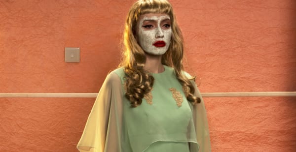 Fashion photography: Woman with a cream facial mask in green dress in front of bright orange wall.