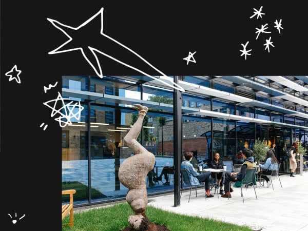 foreground - a sculpture of an upside down person on the grass, background - a group of people sitting at outdoor tables