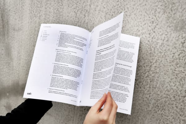 Person's hands holding an open printed white page booklet which has long passages of text. The background is a concrete wall which is in bright natural light.