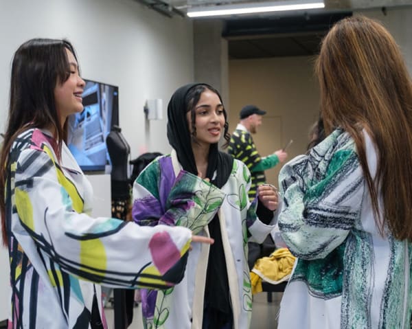 3 students stand together against an orange photoshoot background. They're standing in their printed garments that they designed, talking