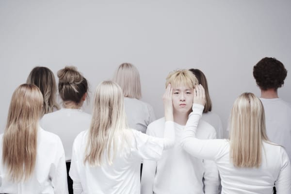 Group of people wearing white tops stood against a plain white wall, 8 of the people are facing the wall so just the back of their heads can be seen. To the right of the central photo one person is facing the camera with 2 of the other people with their hands on the side of the person's head.