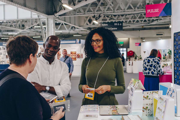 Three people are engaged in a conversation at a booth in a trade show. The booth has various items displayed, and signs for the event and directions are visible in the background.
