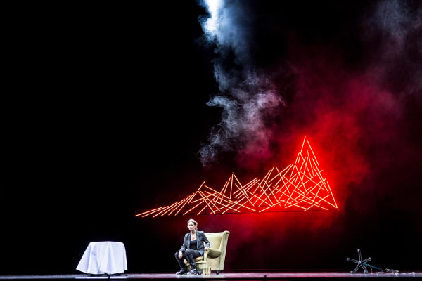 Scene from the opera Happy Happy with Karen Vourc'h. There is a woman sitting on stage in a white armchair with a table. Behind here is red smoke.