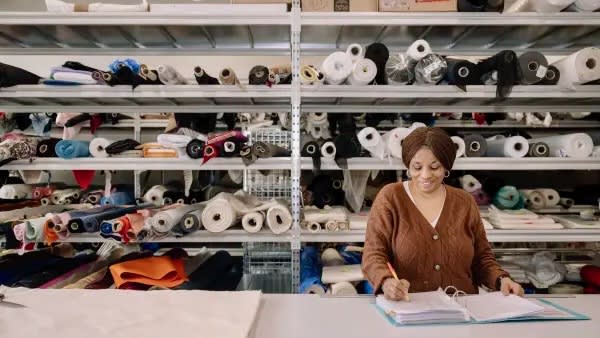 Person writing in a notebook, against a backdrop of shelves with fabrics on.