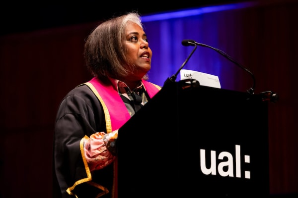 A person stood in front of a lectern mid-way through speaking. The lectern has the UAL logo in white printed on the front and the person is wearing a gold and pink graduation robe. The background is dark and the person is spotlit from above.