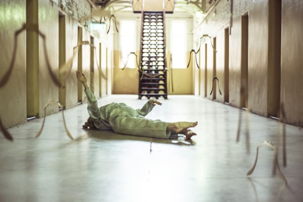  A performer lies prone on the floor, back arched and arms raised in an angular fashion, toes extended. The performer is surrounded by multiple ‘skeletal’ bells, cast in bell bronze, hung at different heights and ritually rung during the performance. The figure lies within a former prison atrium, with multiple openings leading to cells, and an open tread staircase connecting to a mezzanine level, with bright light entering from tall windows to the rear.