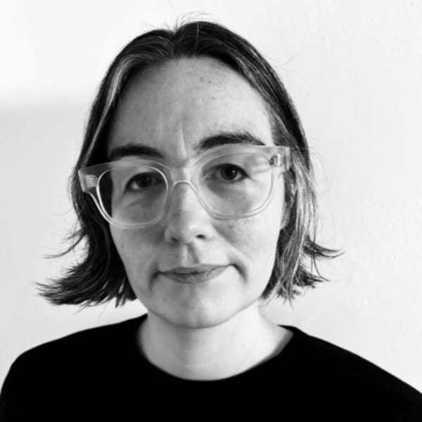 Sarah Lynch, Operations Manager, is standing in front of a white wall wearing a black top, she has clear rimmed glasses and short black hair, the image is black and white. 