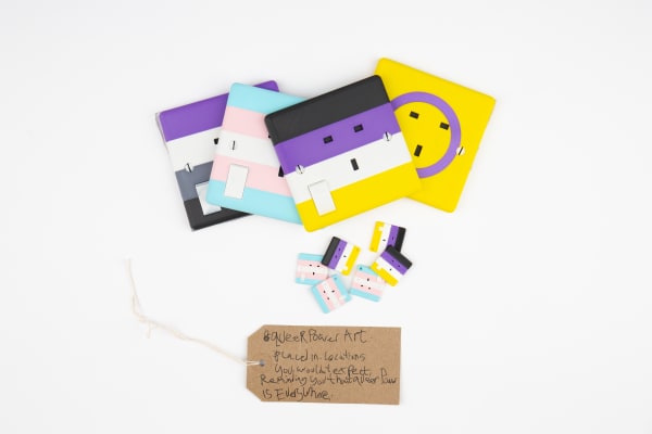 Plug sockets painted in the Trans Pride and Non-binary Pride flagcolours, with a handwritten tag label carrying this caption: 