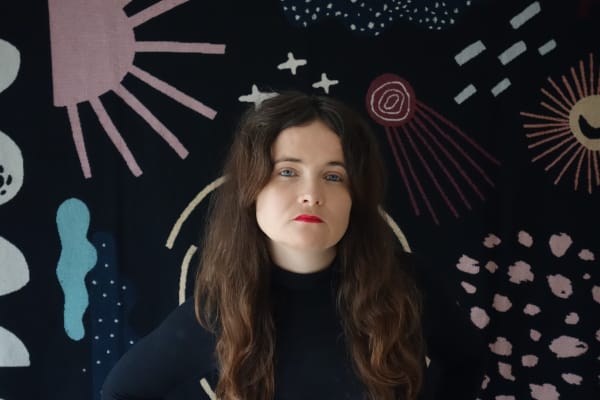 Maya Ober wearing black standing against a patterned background staring at camera.