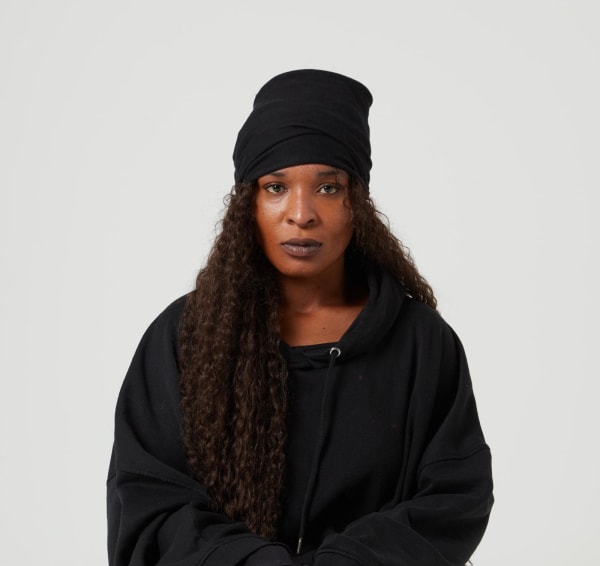 Torishéju Dumi wearing balck, standing against a white backdrop staring at camera.
