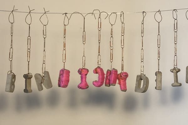 Small concrete letters hanging on a line, dangling on strings of paperclips. Some letters wrapped in pink thread.