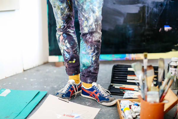 Photo fo legs and feet with paint covered jeans standing among paint brushes and papers