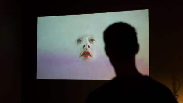 Film projection of a mans face wearing makeup coming out of white and pink mist.