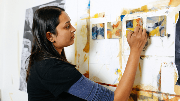 Close up of a student working on 4 small canvases. Paintings are in muted tones with a blue accent.