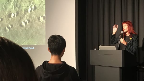 A lecturer speaking into a microphone in front of a slide on a screen
