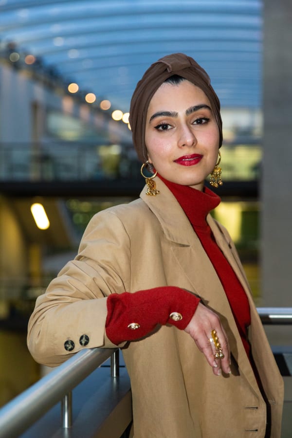 Samar leaning against a railing. She's wearing a headscarf and red lipstick, with a taupe blazer over a dark red roll neck jumper