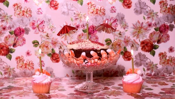 A photo of pink Seafood and cupcakes with golden candles  and prawns festively arranged on a pink  floral background