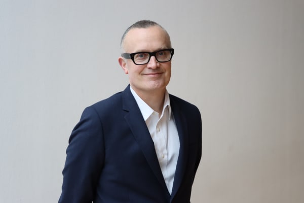 Ben Terrett stands in front of a white wall. He has short, cropped hair, wears dark framed glasses, a white shirt and dark blue suit.