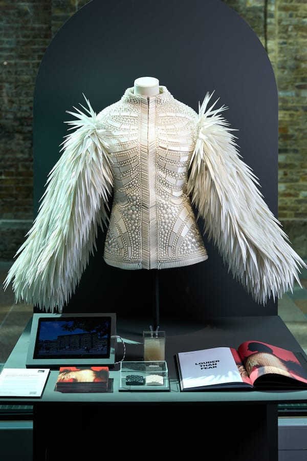 A white coat with a centre seam and small collar, spiky arms and scale-like textures. It is on a table above literature and materials.