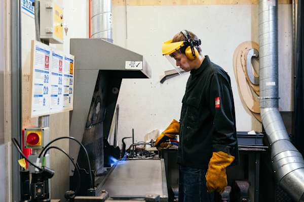 A student in boilersuit and safety helmet, gloves and goggles is operating welding machinery
