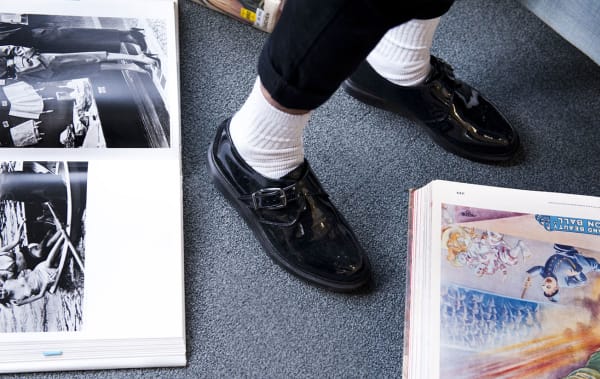 Photo of someone's feet wearing black shiny shoes with white socks standing among books on the floor