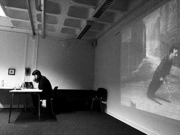 black and white photo of person sitting at a desk in the corner of a classroom. Behind them a large projector shows a still image of a Charlie Chaplin skit.