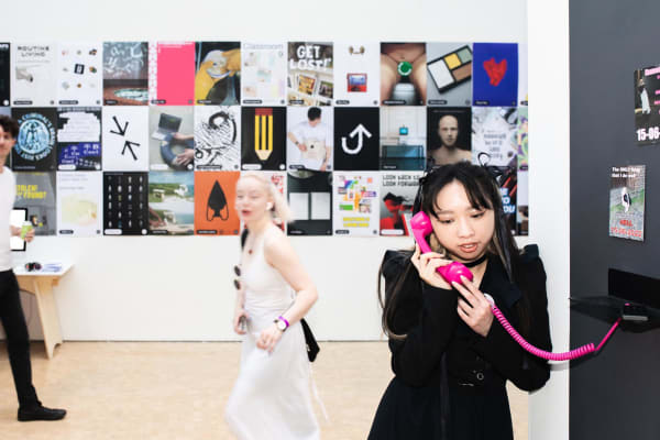 Figure holding pink telephone standing in front of posters on white walll
