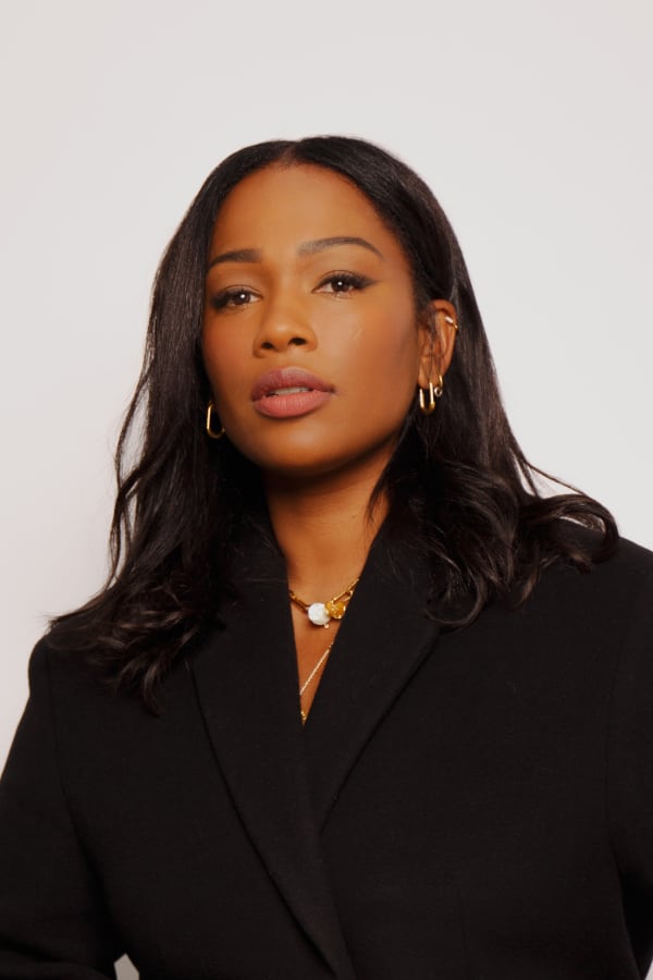 Sharmadean Reid is in front of a grey background looking slightly down at the camera. Sharmadean has long, dark hair and wears a black jacket.