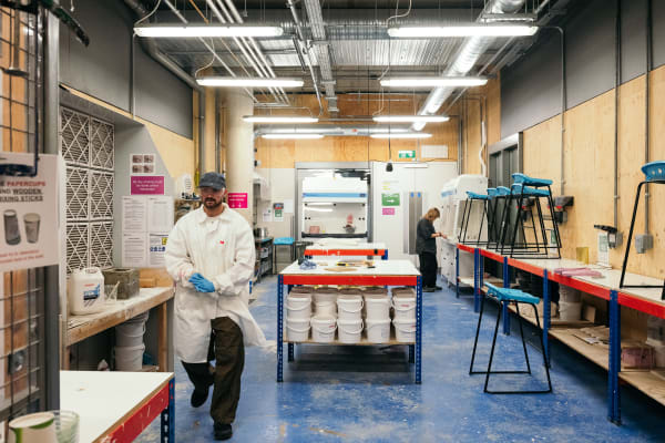 A wide shot of the casting workshop with a central island storing white buckets and a long-tabletop. At the far end a student is working at a large machine. Toward the left of the image, a technician in white lab coat, blue gloves and safety goggles is walking toward the camera.