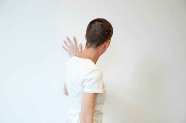 A person wearing a white t-shirt facing a white wall and touching it