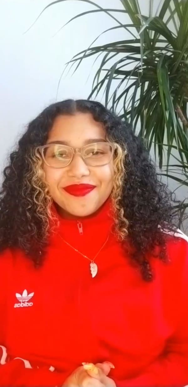 Head shot of smiling dark haired female with curly hair, glasses in a red top and red lipcolour with a green plant in the backround