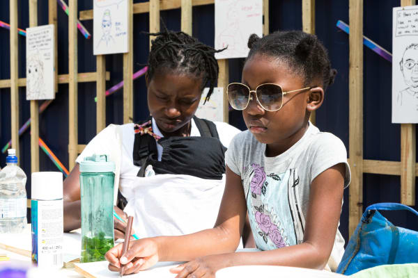 A woman with a baby in a pouch and a little girl wearing sunglasses drawing against a shipping container background