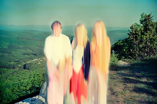 Photograph of 3 people with blurred out faces standing at top of a mountain with lush green forests below and clear blur sky.