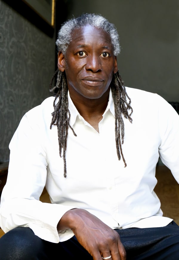 Trevor Robinson sits in front of patterned, wallpapered wall and looks directly into camera. He has long, dark greying hair and wears a white shirt and dark blue trousers.