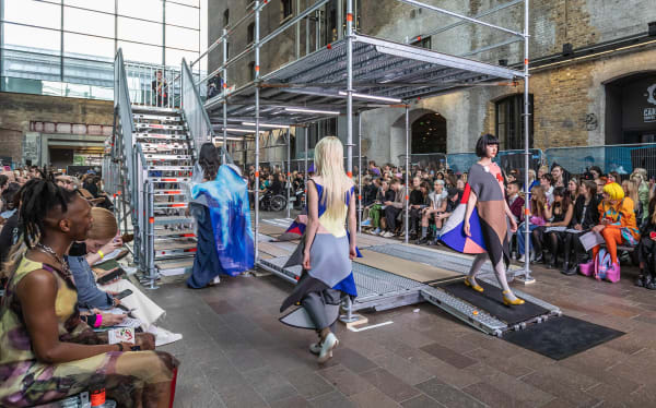 Two floor catwalk made in scaffold with three models wearing colourful garments