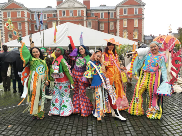 An image of 6 people wearing  bright, colourful outfits. Each person's outfit is unique and full of patterns, textures and messages. They're stood in front of the crows at Chelsea Parade Ground and posing and smiling at the camera. 