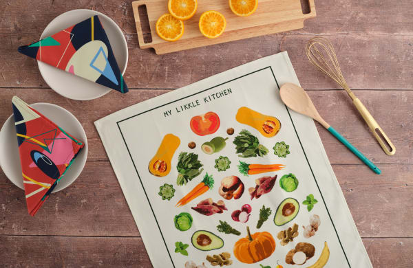 A dark wooden table takes up the whole image. At the very top of the image there is a wooden cutting board with cut orange slices. To the left of the image there are two bowls, each with a colourful retro print napkin in the centre. The middle and bottom right of the image feature a poster printed with various fruits and vegetables, and the top of the poster reads “My Likkle Kitchen”. On the right next to the poster there is a wooden spoon and a gold whisk.