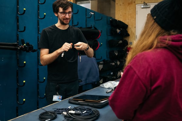 A student getting support at the Central Loan Store. A staff member behind the counter is holding a cable and talking. Behind him are tall cabinets with some equipment visible.