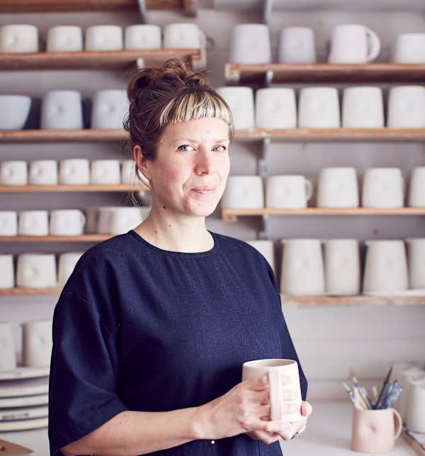 A colour portrait photo of Emma in the ceramics studio. Behind her are rows of mugs.