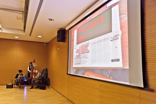 A projector screen, with someone playing a cello to the side. 