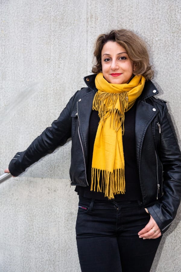 Halime stood in front of a concrete wall. She's wearing a black leather jacket, a yellow scarf and red lipstick