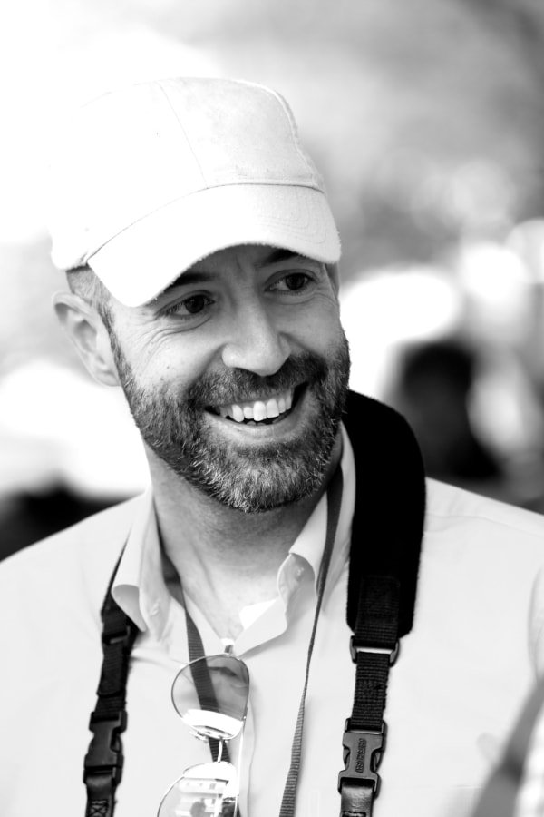 Black and white portrait photo of Giulio. He's wearing a cap, a polo shirt and has a camera strap round his neck. He's smiling.