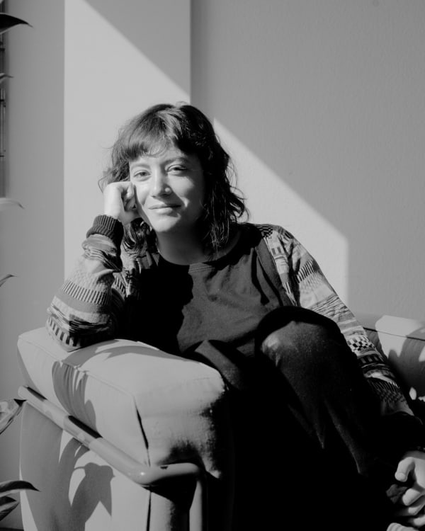 Black and white shot of Nina Paim. Nina sits on a sofa with legs curled up. Shew has long hair and wears a a patterned cardigan. 