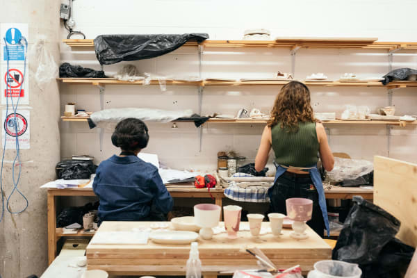 Students standing back to camera at shelves in the ceramics workshop. In the foreground is a countertop with materials for ceramic production and some finished designs.