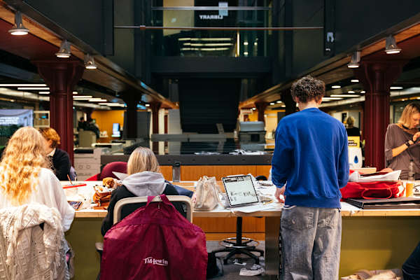 Students standing and seated at a long table in CSM library, with reading materials, bags and laptops visible.