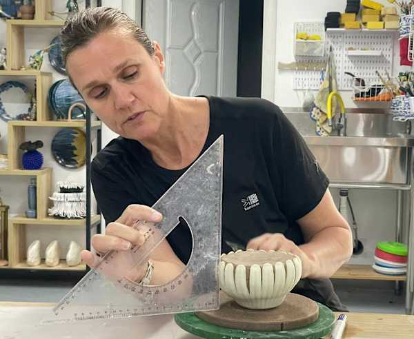 A colour portrait photo of Helen working in the studio. She is holding a set square and working on a design. Behind her are shelves of ceramics and a sink.