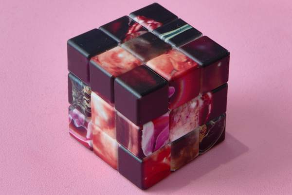 A partially twisted cube puzzle made of glossy black and translucent pink marbled blocks sits on a pink surface.